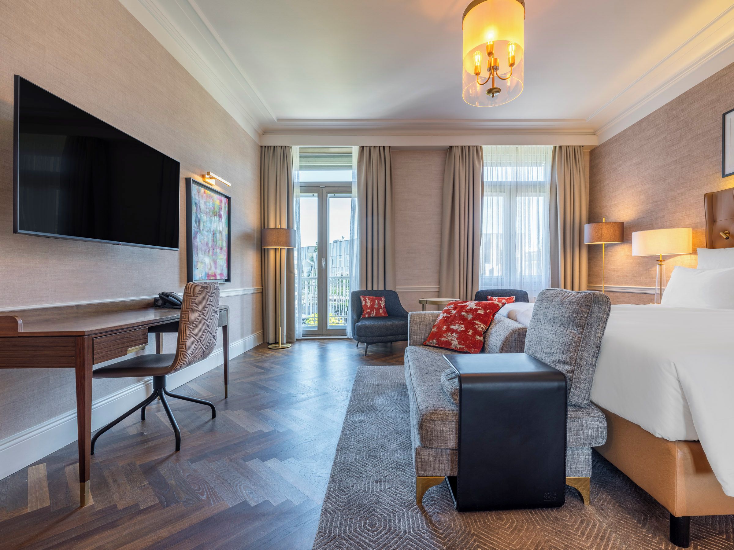 A hotel room with a flat-screen TV, a desk, a sofa, a bed, and a chandelier.