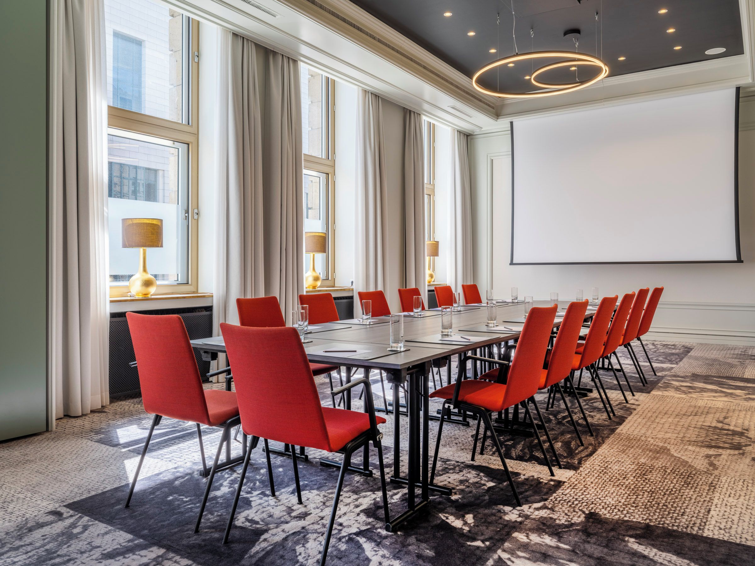 Un salón de conferencias con una larga mesa, sillas rojas, una pantalla de proyector y ventanas amplias con cortinas.
