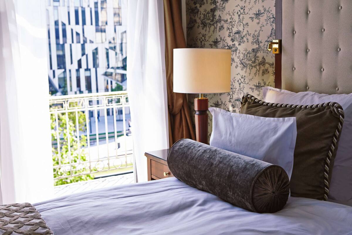 Une chambre d’hôtel avec un lit, une table de chevet, une lampe et une vue sur un immeuble par la fenêtre.