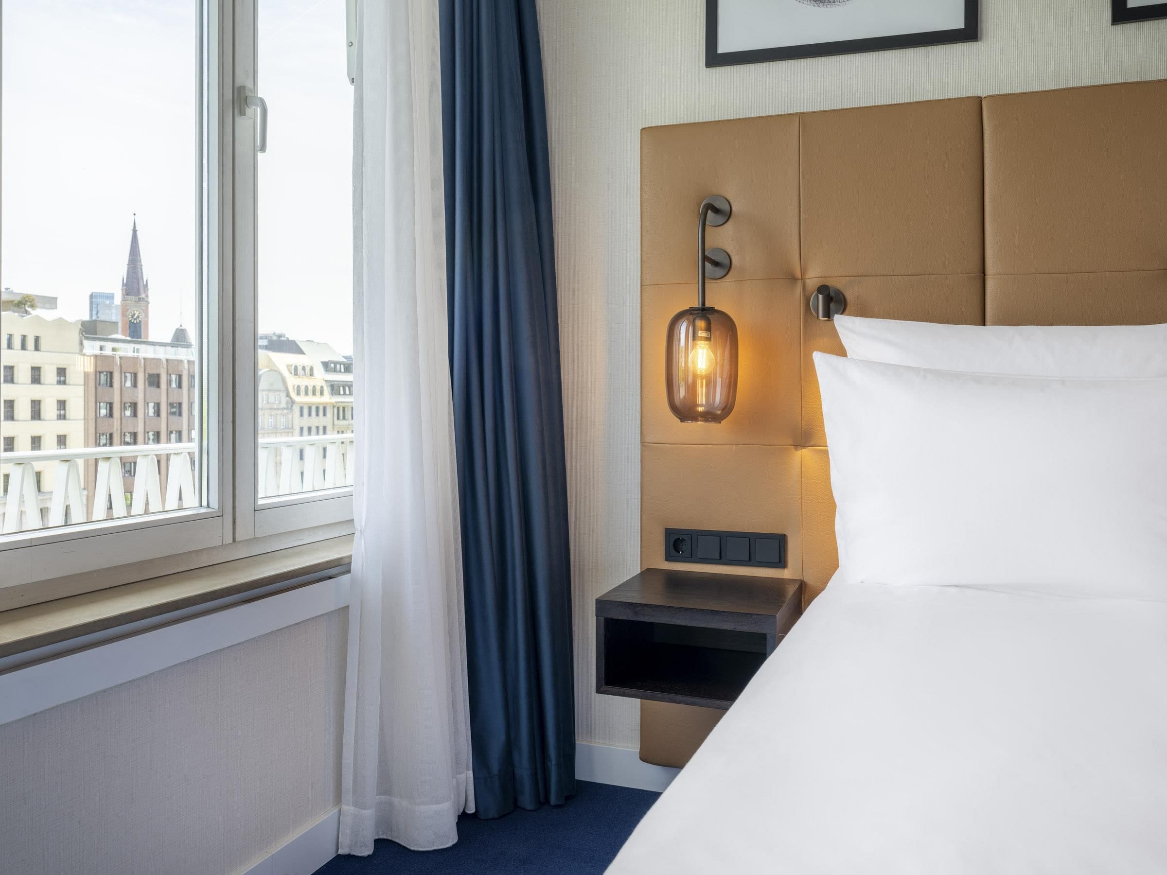 Hotelkamer met een bed, nachtkastje, een wandlamp, raam met gordijnen en uitzicht op gebouwen.