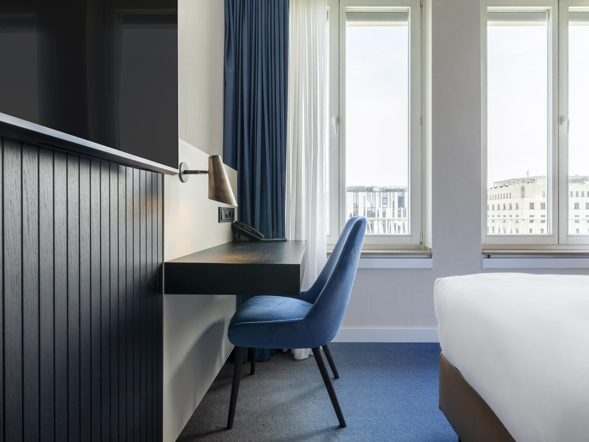 Una camera d'albergo con un letto, una scrivania con una lampada, una sedia e una finestra vista edifici.