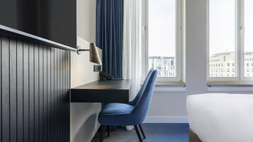 Una camera d'albergo con un letto, una scrivania con una lampada, una sedia e una finestra vista edifici.