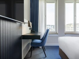 Una camera d'albergo con un letto, una scrivania con una lampada, una sedia e una finestra vista edifici.