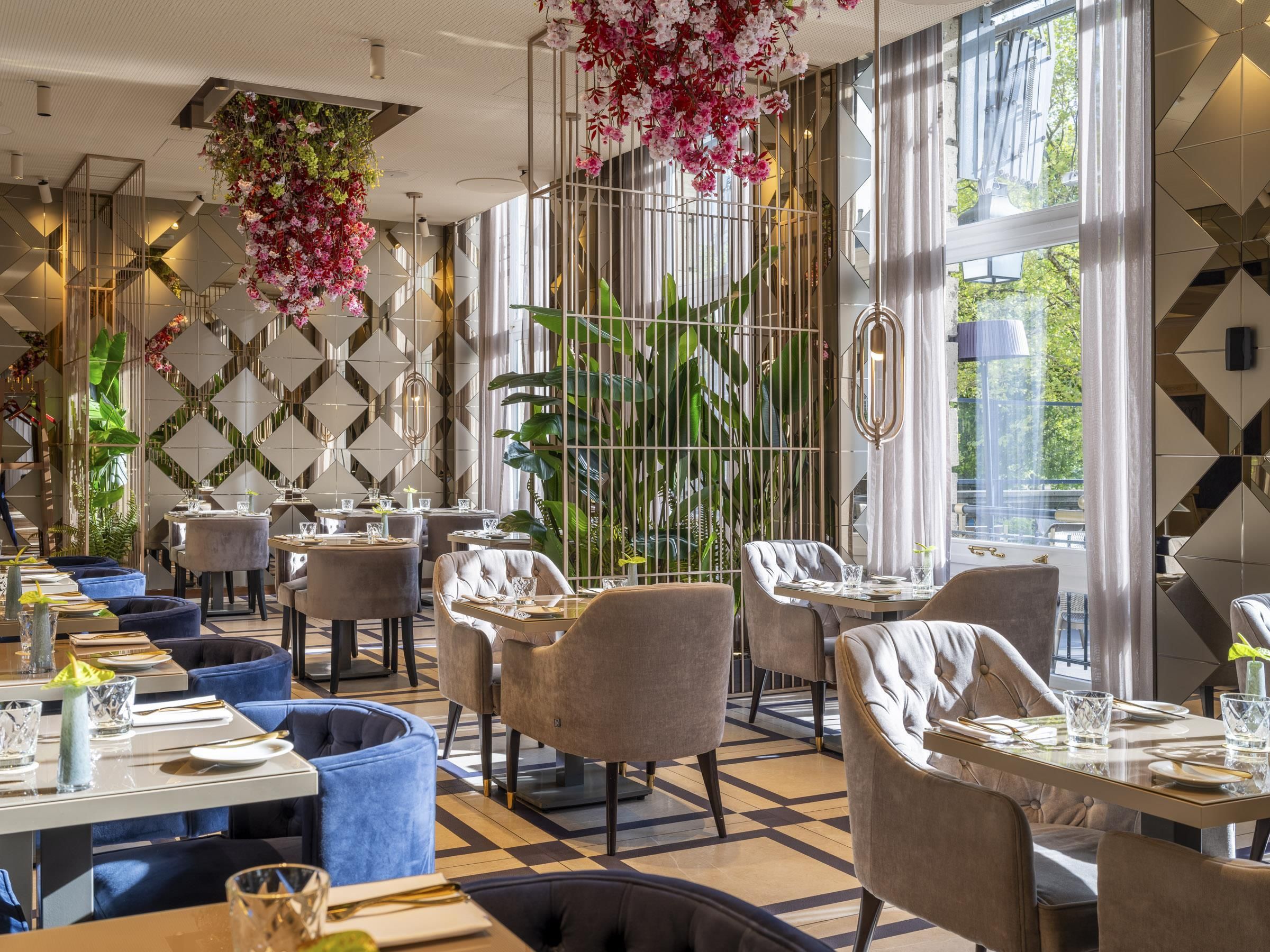 Un restaurante con una decoración moderna, que presenta patrones geométricos en las paredes, arreglos florales colgantes y una variedad de opciones de asientos, incluyendo mesas redondas y sillas con cojines.