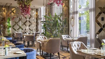 Un restaurante con una decoración moderna, que presenta patrones geométricos en las paredes, arreglos florales colgantes y una variedad de opciones de asientos, incluyendo mesas redondas y sillas con cojines.