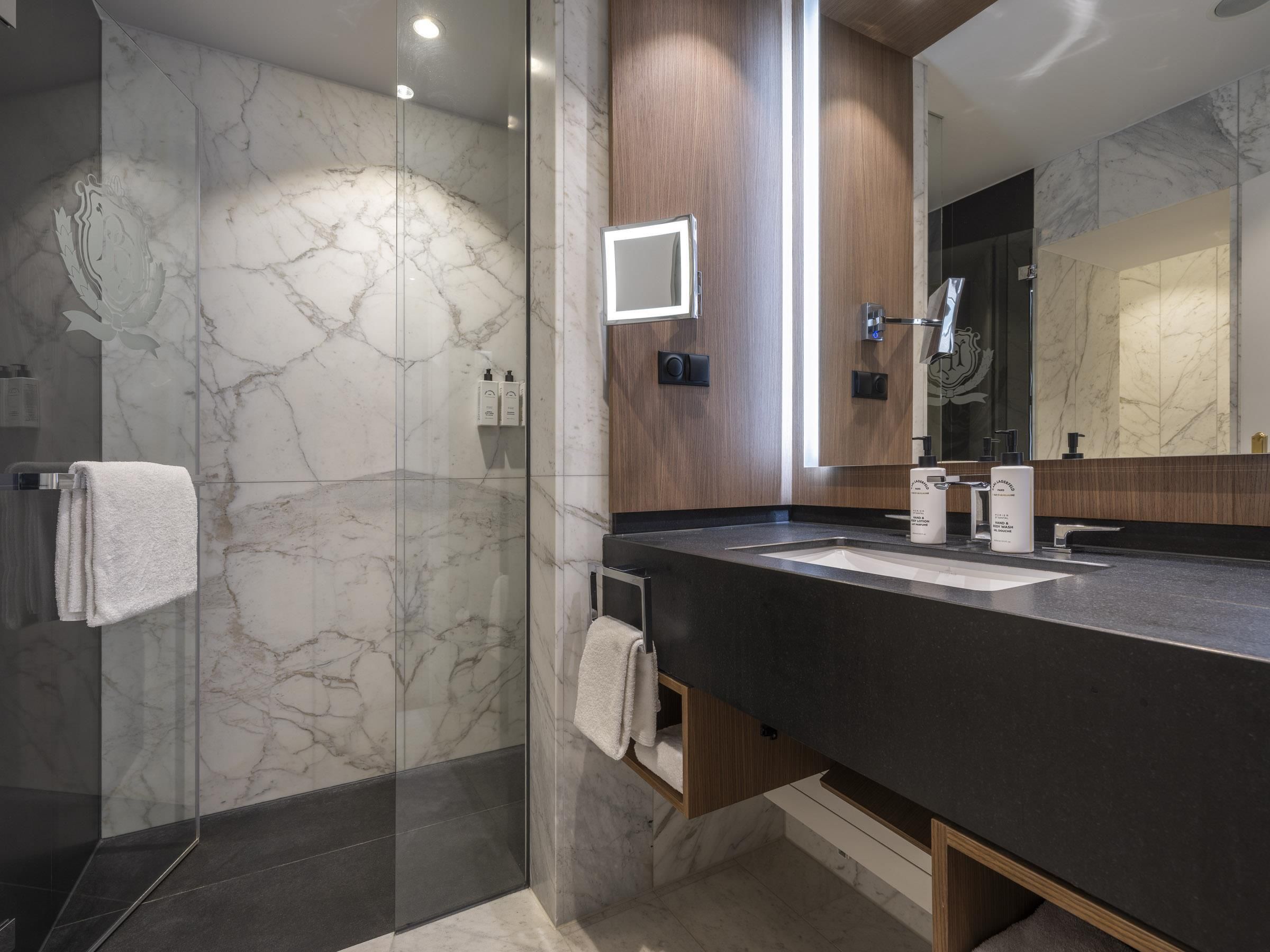 A modern bathroom with a glass shower enclosure, marble walls, a dark countertop, and a large mirror.