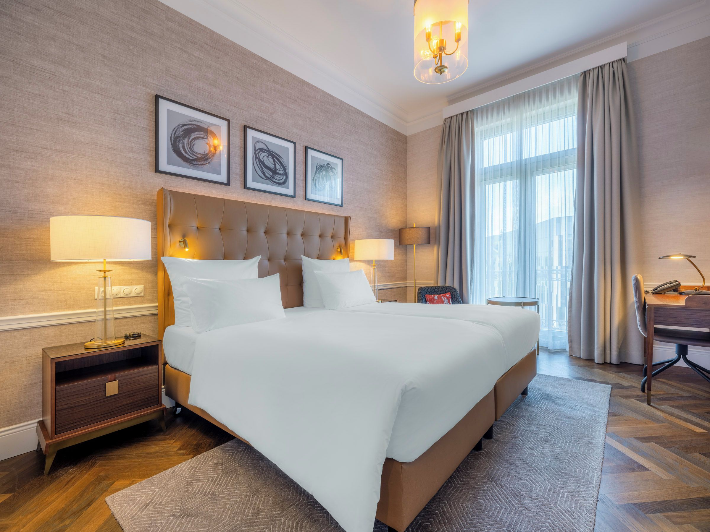 A hotel room with a large bed, two bedside tables, a desk, and a chair. There are three framed artworks on the wall, a chandelier, and a large window with curtains.