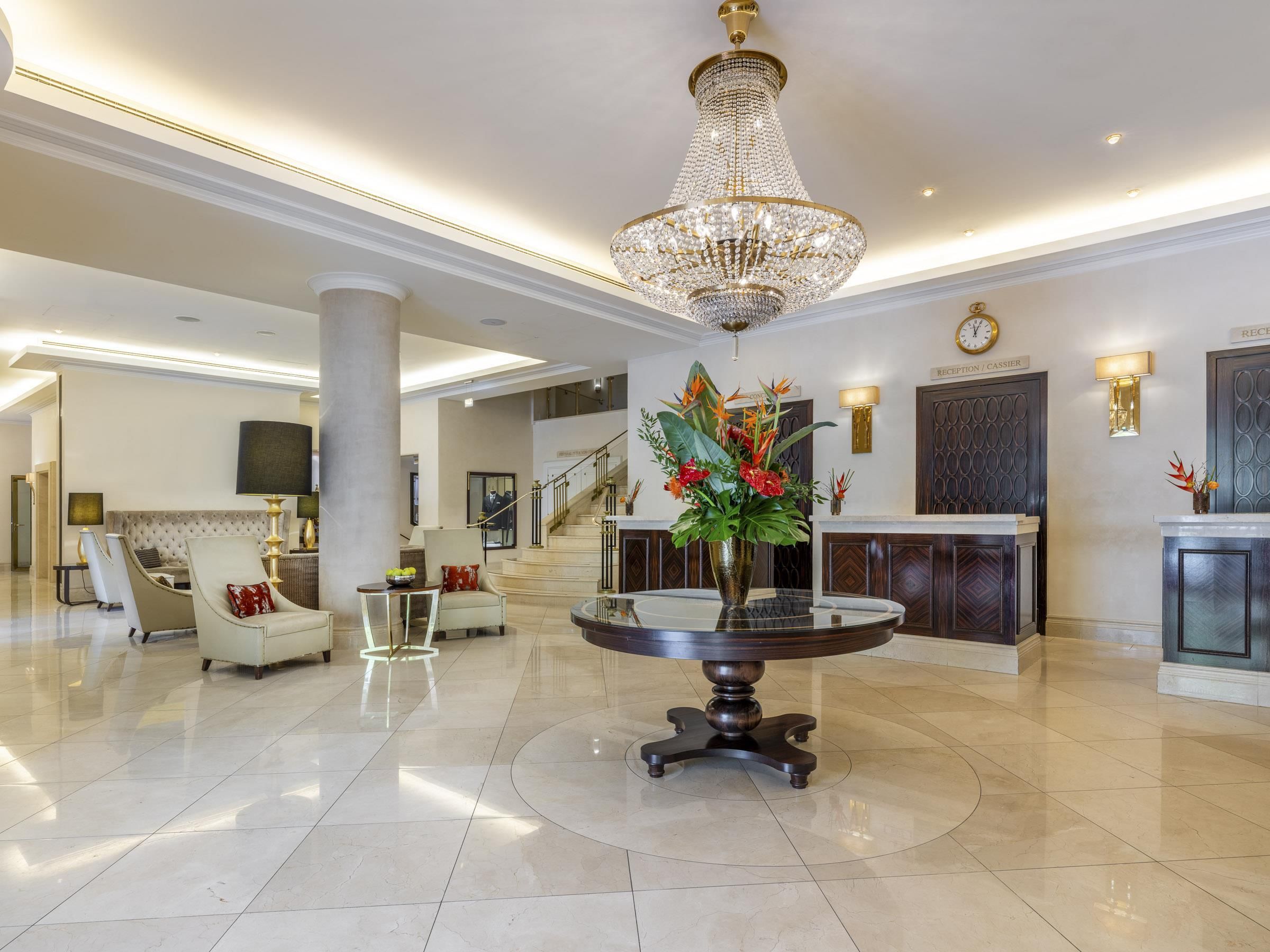 A spacious lobby with a grand chandelier, a reception desk, and a staircase leading to upper floors.