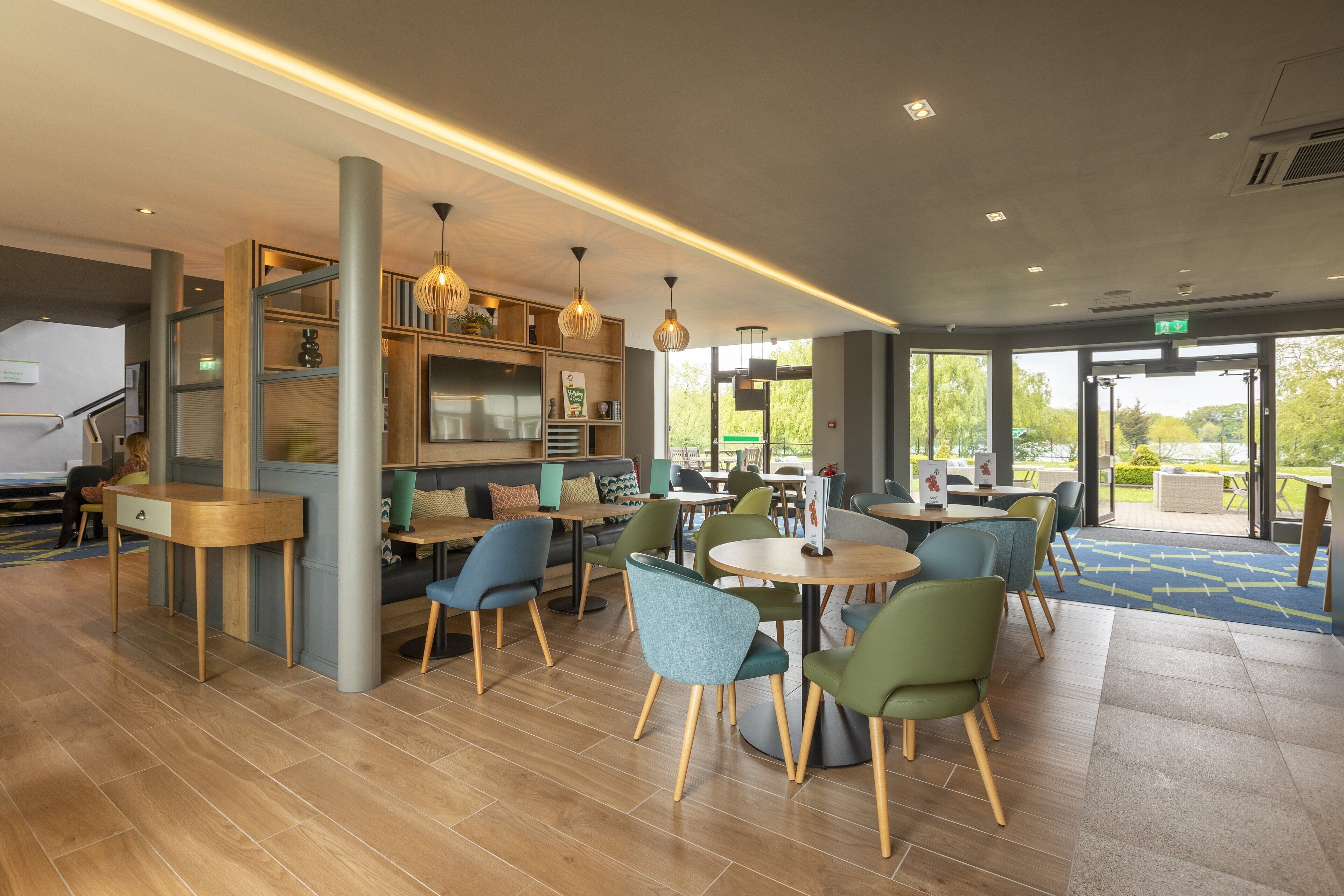 A modern indoor space with wooden flooring, green and blue chairs, and a variety of tables. There are shelves with items and pendant lights.