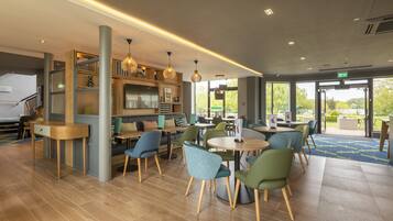 A modern indoor space with wooden flooring, green and blue chairs, and a variety of tables. There are shelves with items and pendant lights.