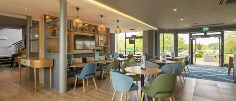 A modern indoor space with wooden flooring, green and blue chairs, and a variety of tables. There are shelves with items and pendant lights.