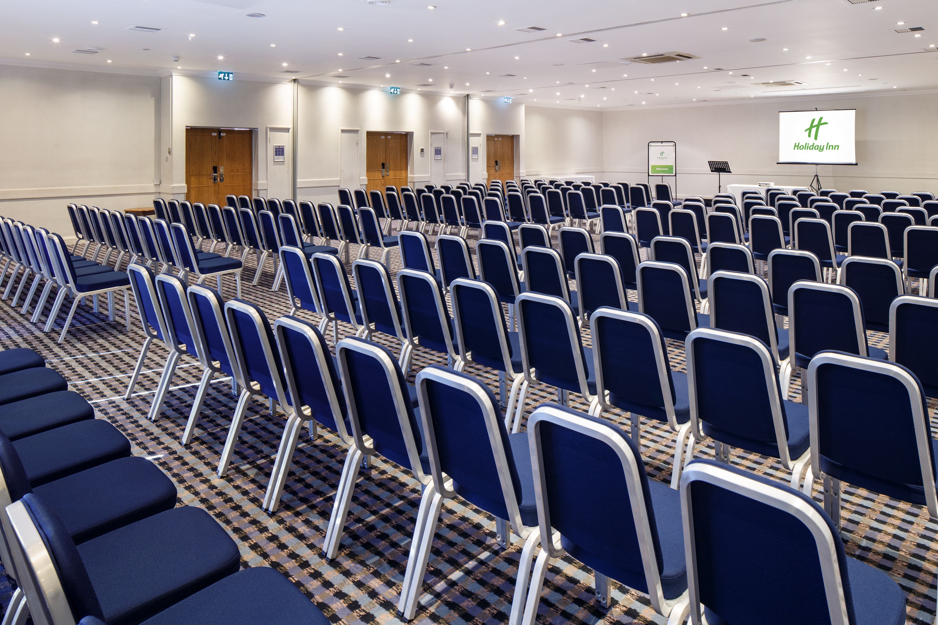 A conference room with rows of blue chairs, a carpeted floor, and a screen displaying the Holiday Inn logo.