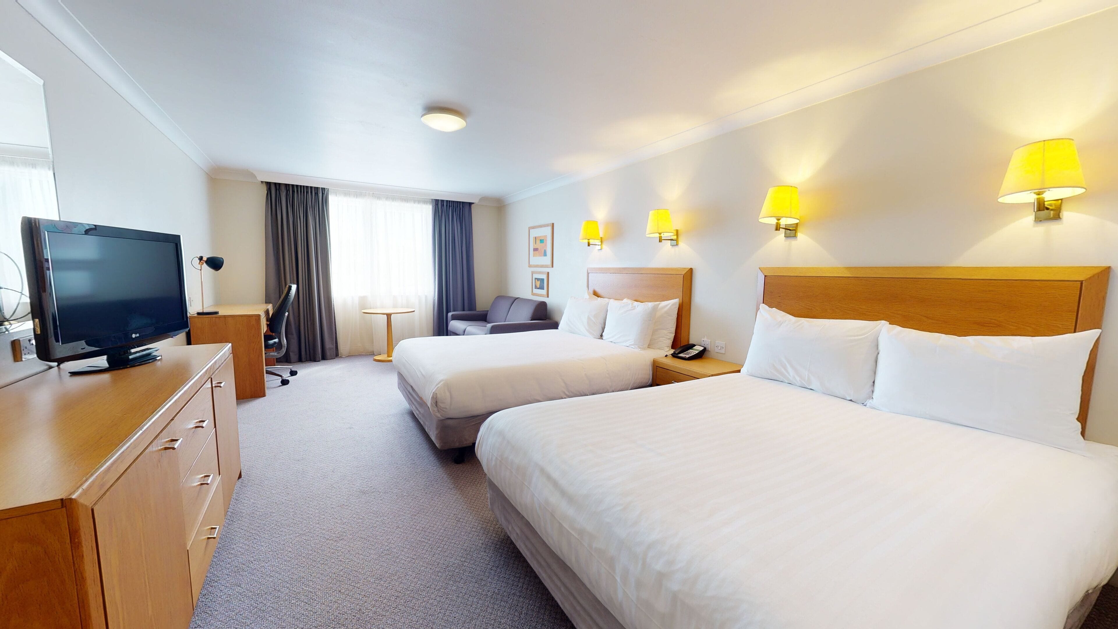 A hotel room with two beds, a desk with a TV, a chair, a small table, and wall-mounted lamps.
