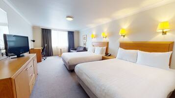 A hotel room with two beds, a desk with a TV, a chair, a small table, and wall-mounted lamps.