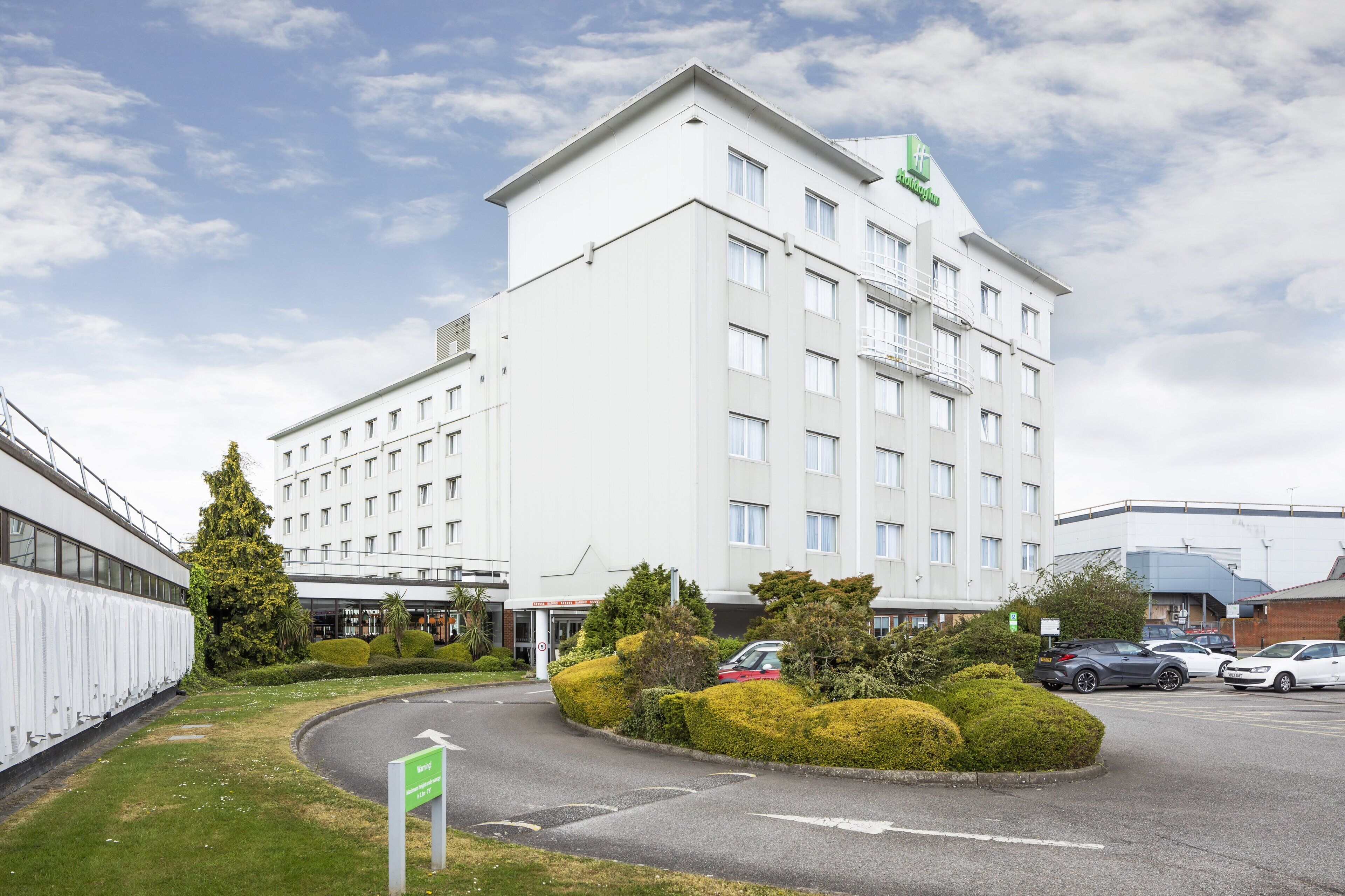 A modern hotel with a white exterior, multiple floors, and a parking area with several cars.