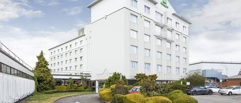 Un hôtel moderne à la façade blanche, avec de nombreux étages et un parking où sont stationnées plusieurs voitures.