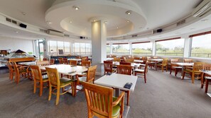 Un restaurante amplio con varias mesas y sillas, ventanas grandes que ofrecen vistas, y una columna central.