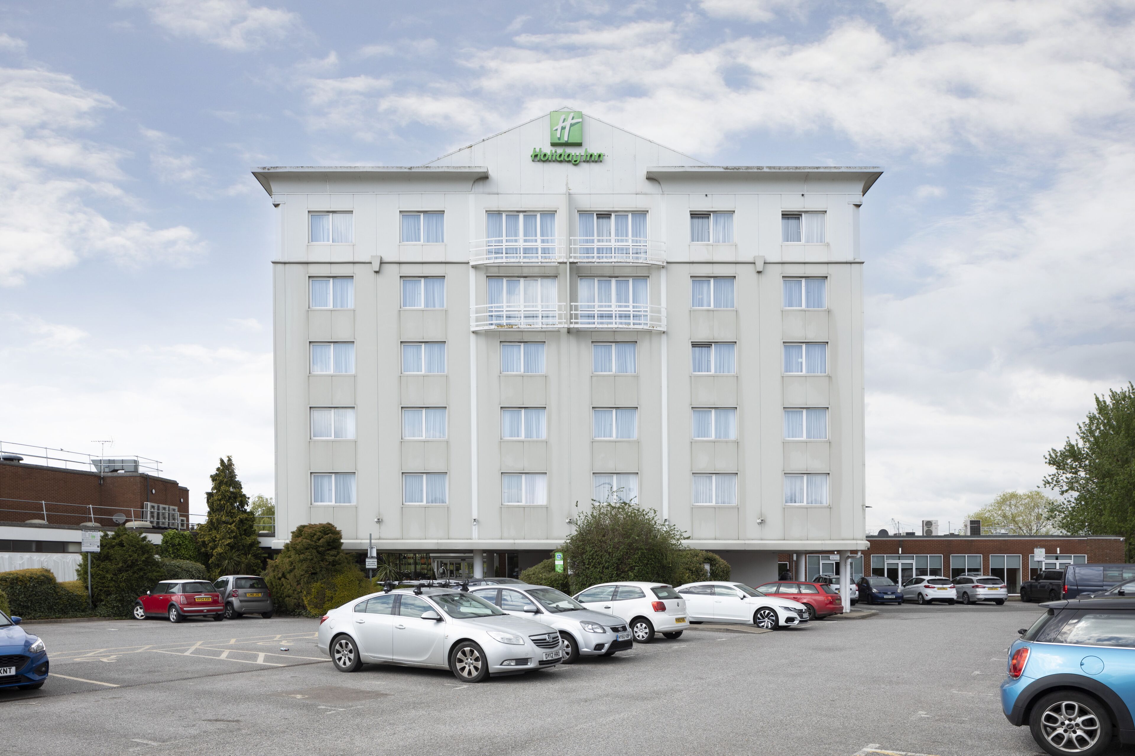 A large, multi-story hotel with a prominent green logo, several balconies, and a parking lot with various cars.