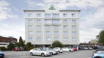 A large, multi-story hotel with a prominent green logo, several balconies, and a parking lot with various cars.