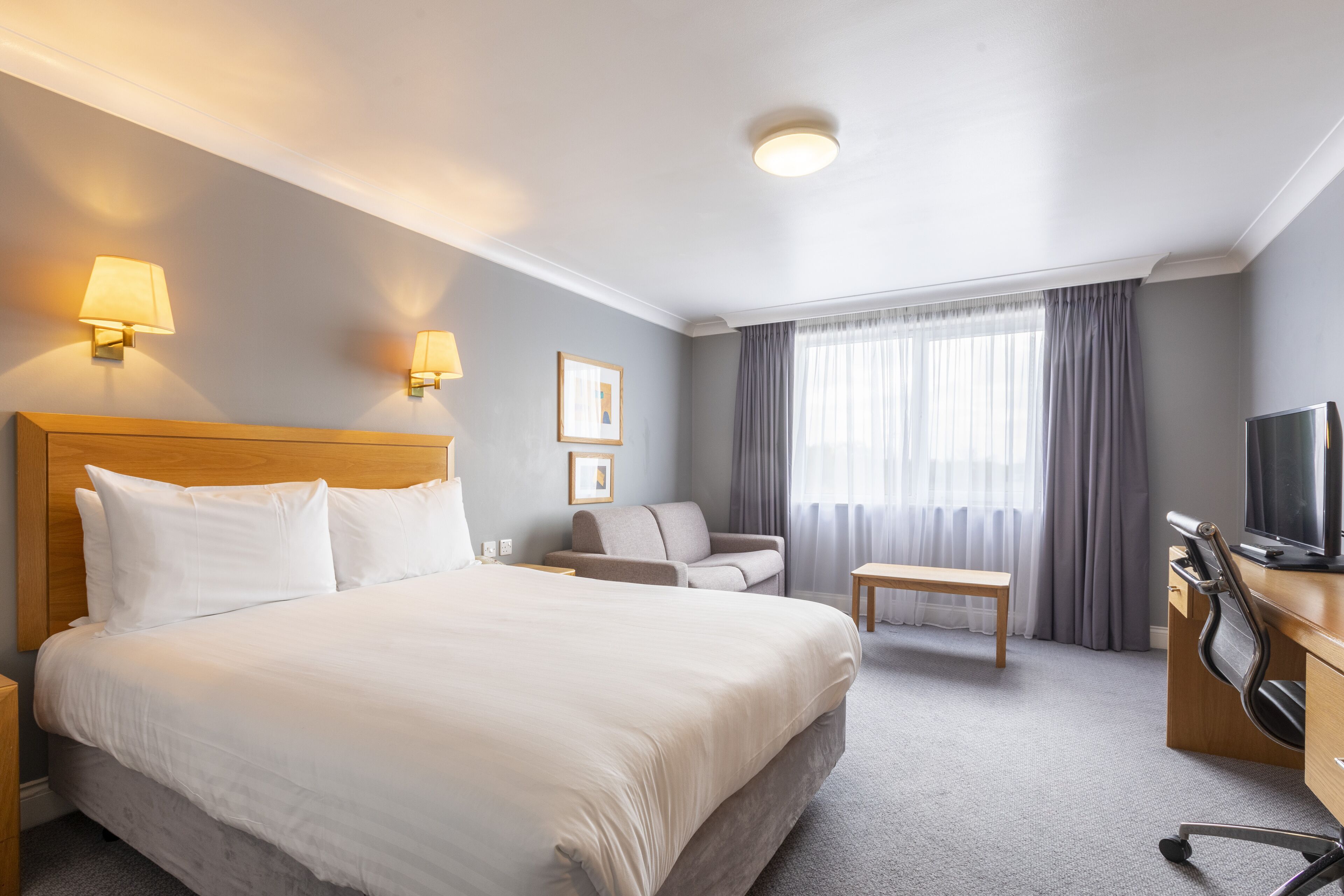 A hotel room with a large bed, a desk with a chair, a television, and two wall-mounted lamps.