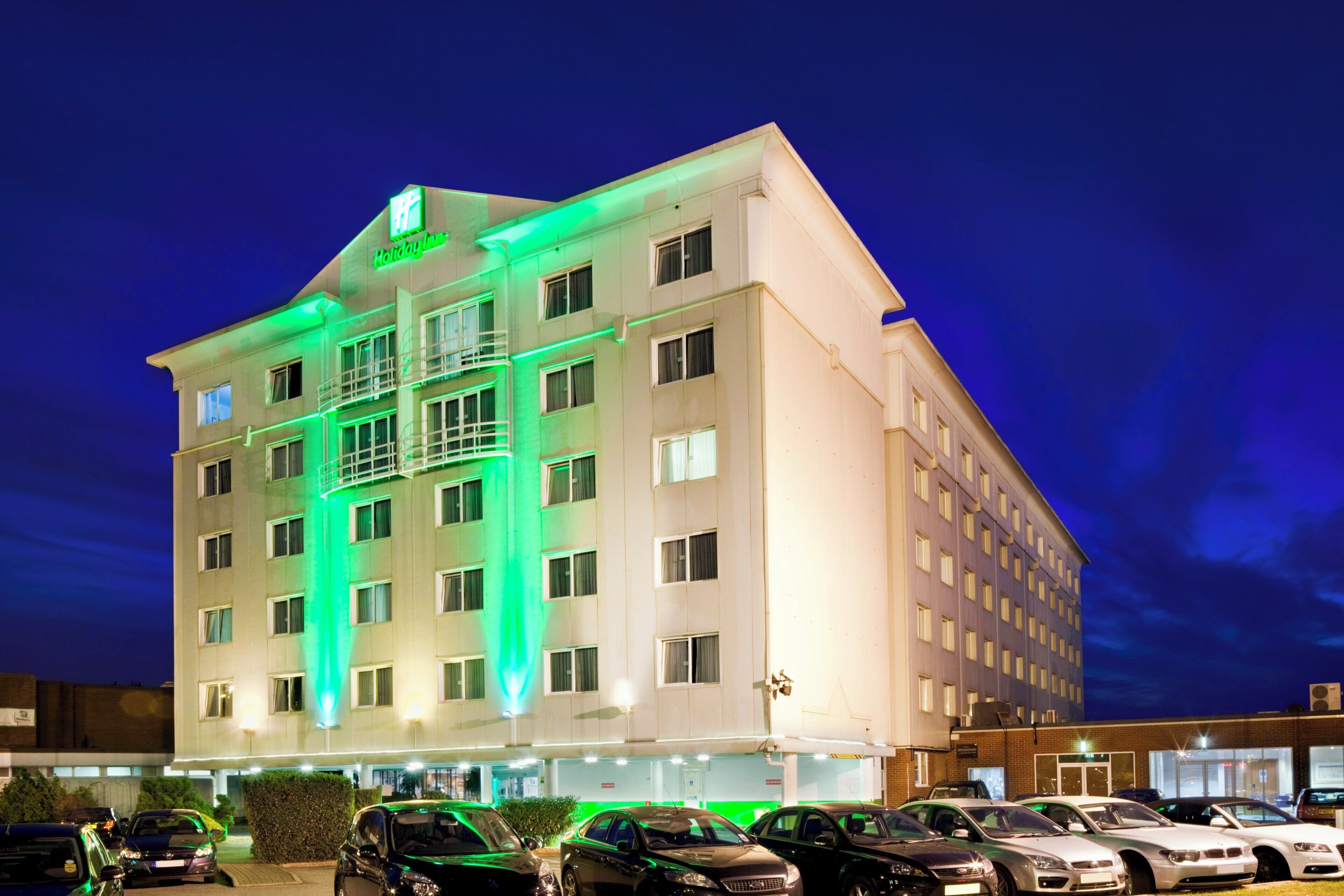A multi-story hotel with a prominent 'Holiday Inn' sign, a parking lot with several cars, and a clear night sky.