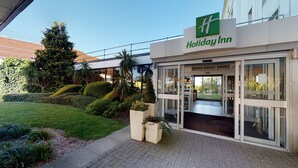 Entrada de un hotel Holiday Inn con puerta de vidrio transparente, césped cuidado y diversas plantas.