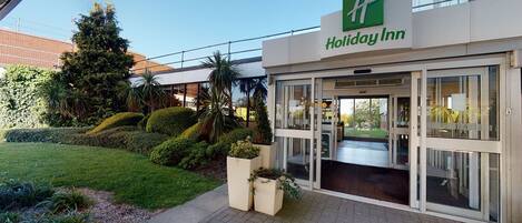 A Holiday Inn hotel entrance with a clear glass door, a well-maintained lawn, and a variety of plants.