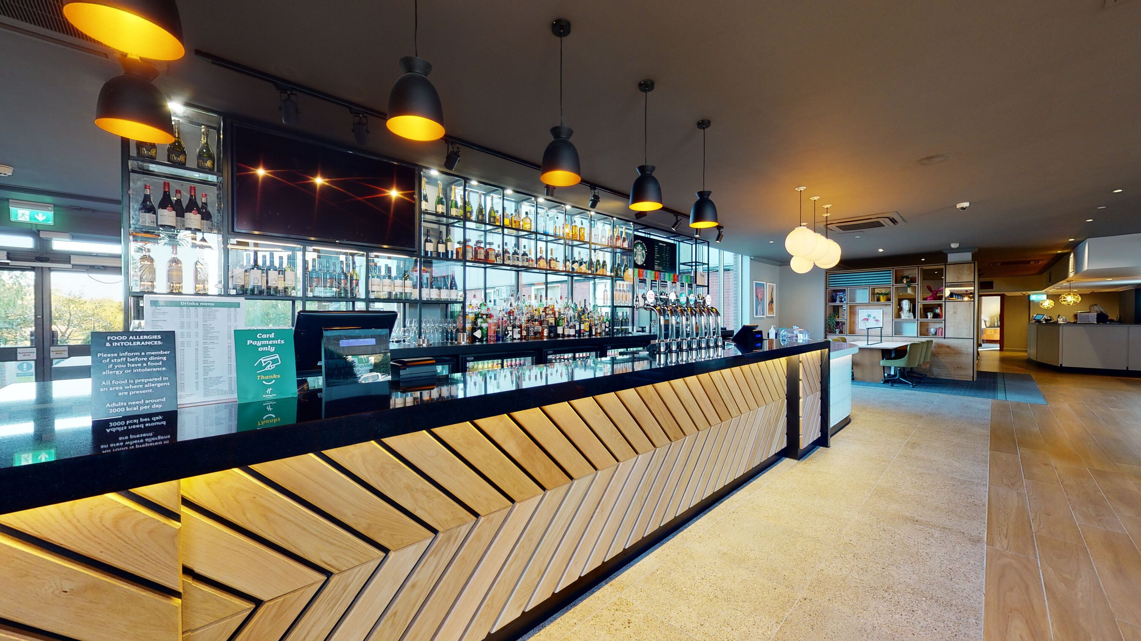 Zona de bar con mostrador de madera, estantes con botellas e iluminación colgante.