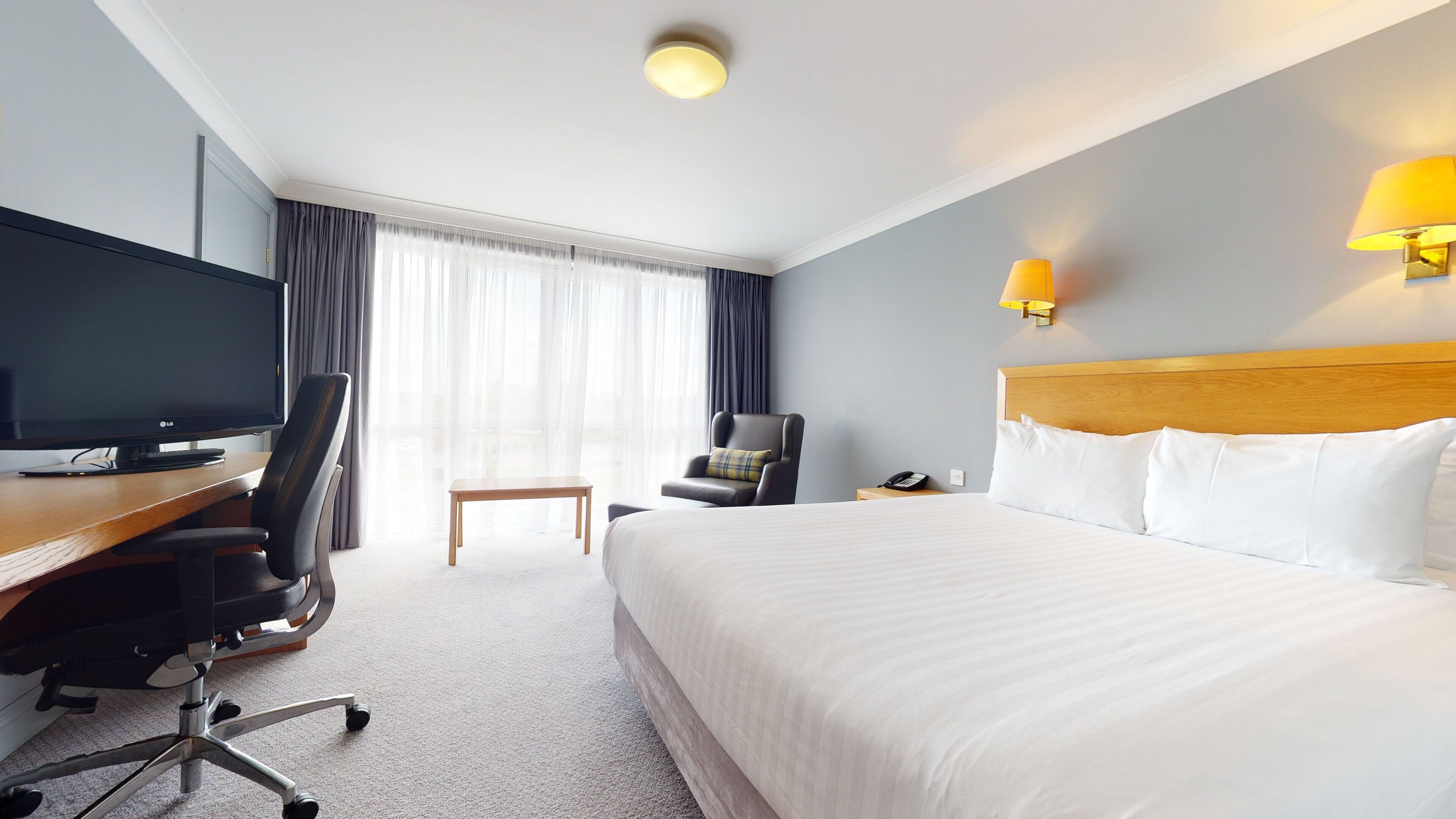 Et hotelværelse med seng, skrivebord med tv, stol, lampe og et vindue med gardiner.
