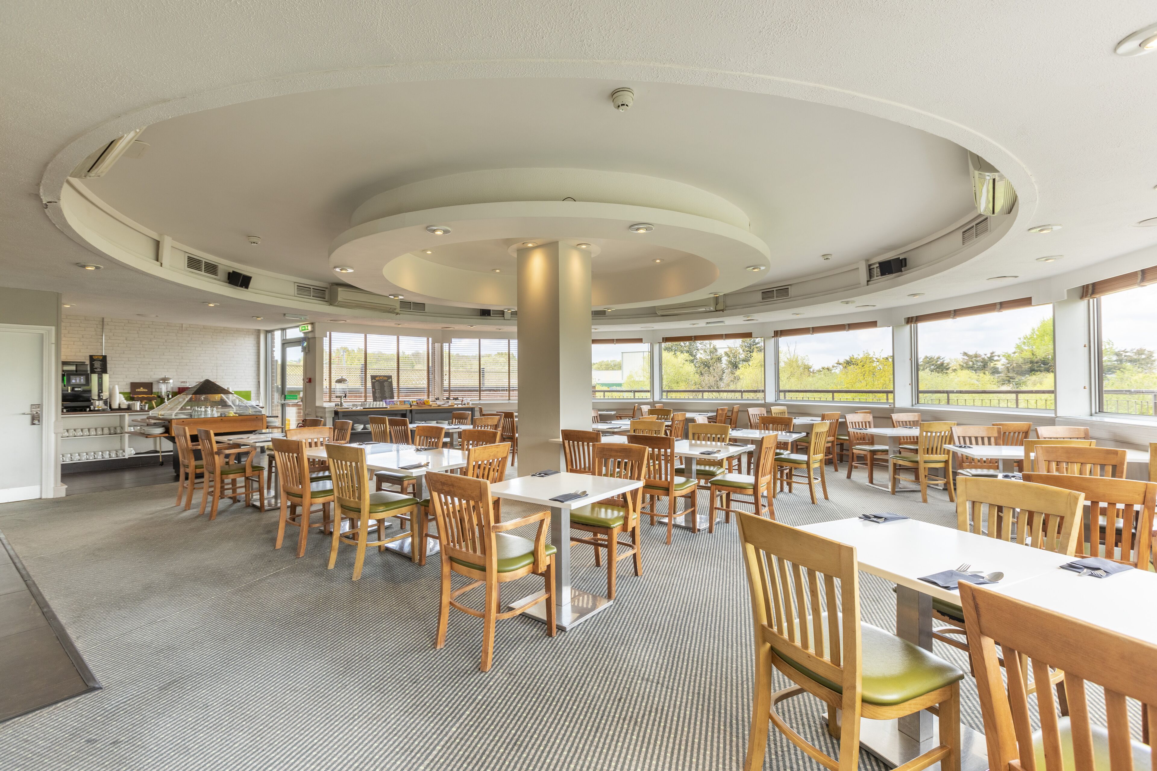 A spacious dining area with multiple tables and chairs, large windows offering views of the outdoors, and a modern ceiling design.