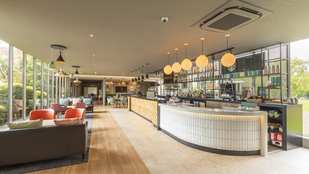 Un moderno bar con una barra curva, asientos con cojines coloridos y grandes ventanales que ofrecen vistas al verde.