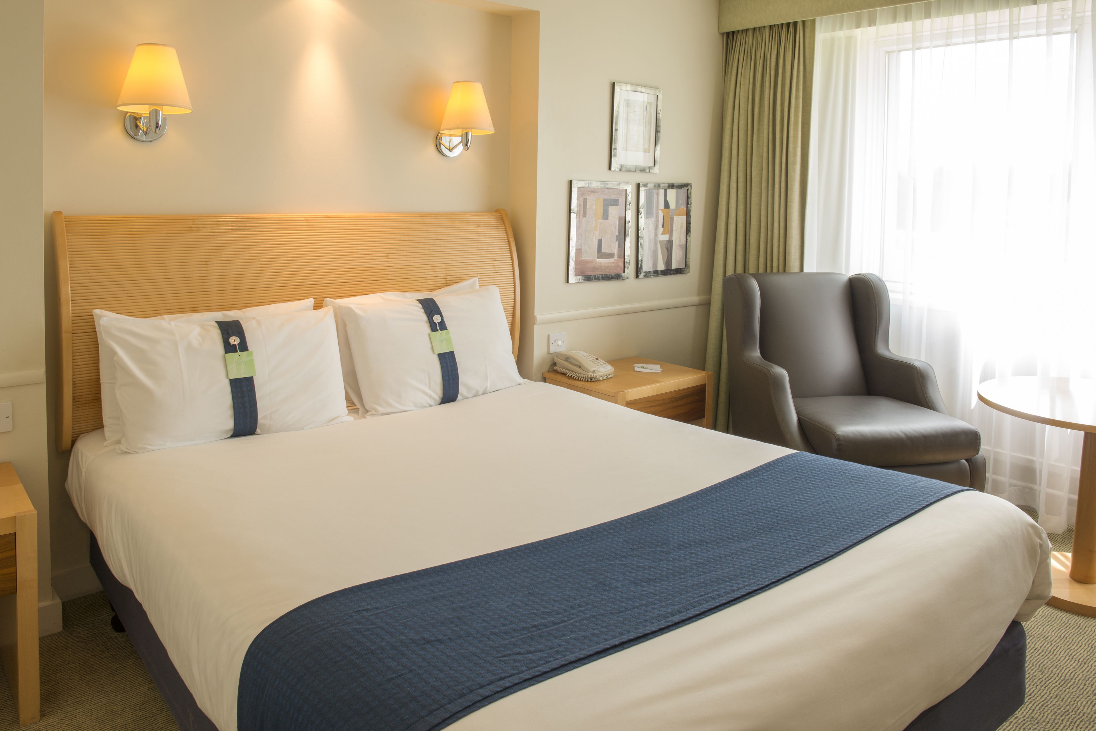 Habitación de hotel con una cama grande, dos lámparas de noche, una silla, una mesita y un teléfono en la mesita de noche.