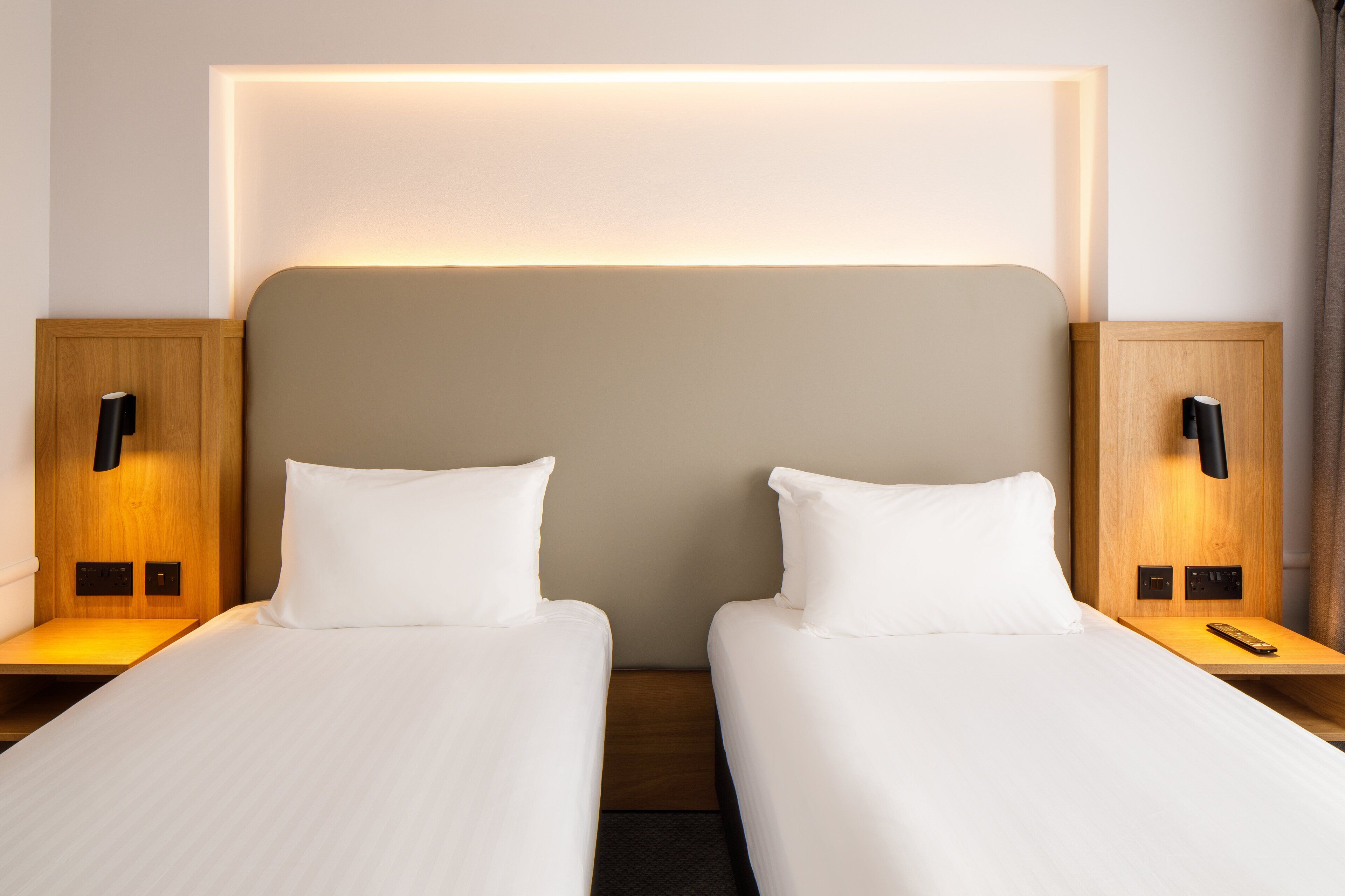 Habitación de hotel con dos camas individuales, cabecero de madera, mesitas de noche con lámparas y una lámpara moderna sobre la cama.