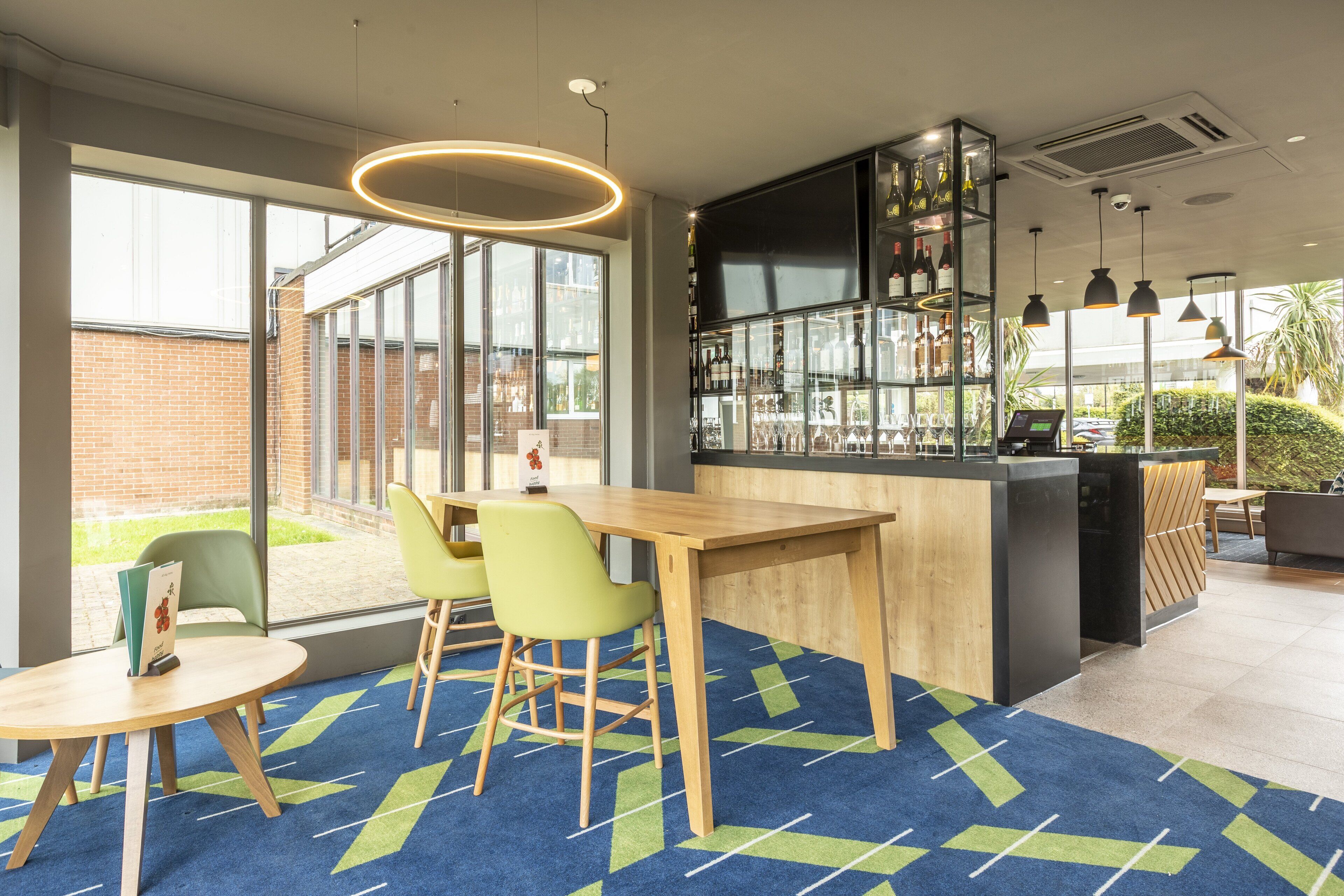 A modern bar area with a wooden counter, green chairs, and a glass display of bottles.