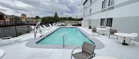 A swimming pool with lounge chairs and tables, surrounded by a fence and buildings.