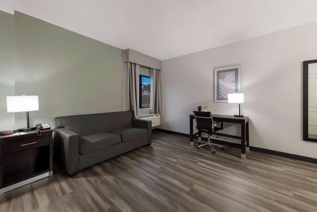 A hotel room with a grey sofa, a desk with a chair, a lamp, and a window with curtains.