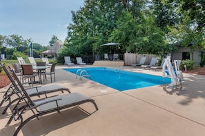 Piscina com espreguiçadeiras e mesas, cercada por árvores e um muro.