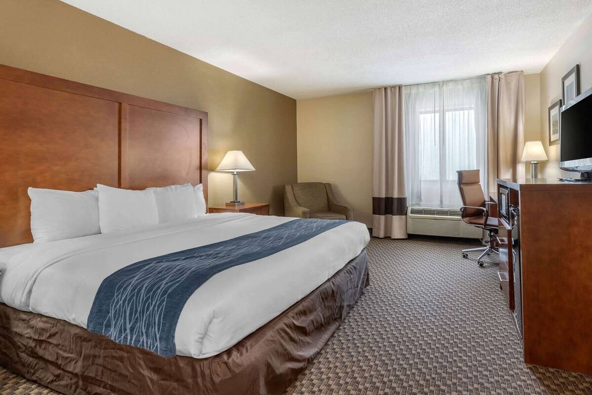 A hotel room with a large bed, a desk with a television, a chair, and a desk lamp.