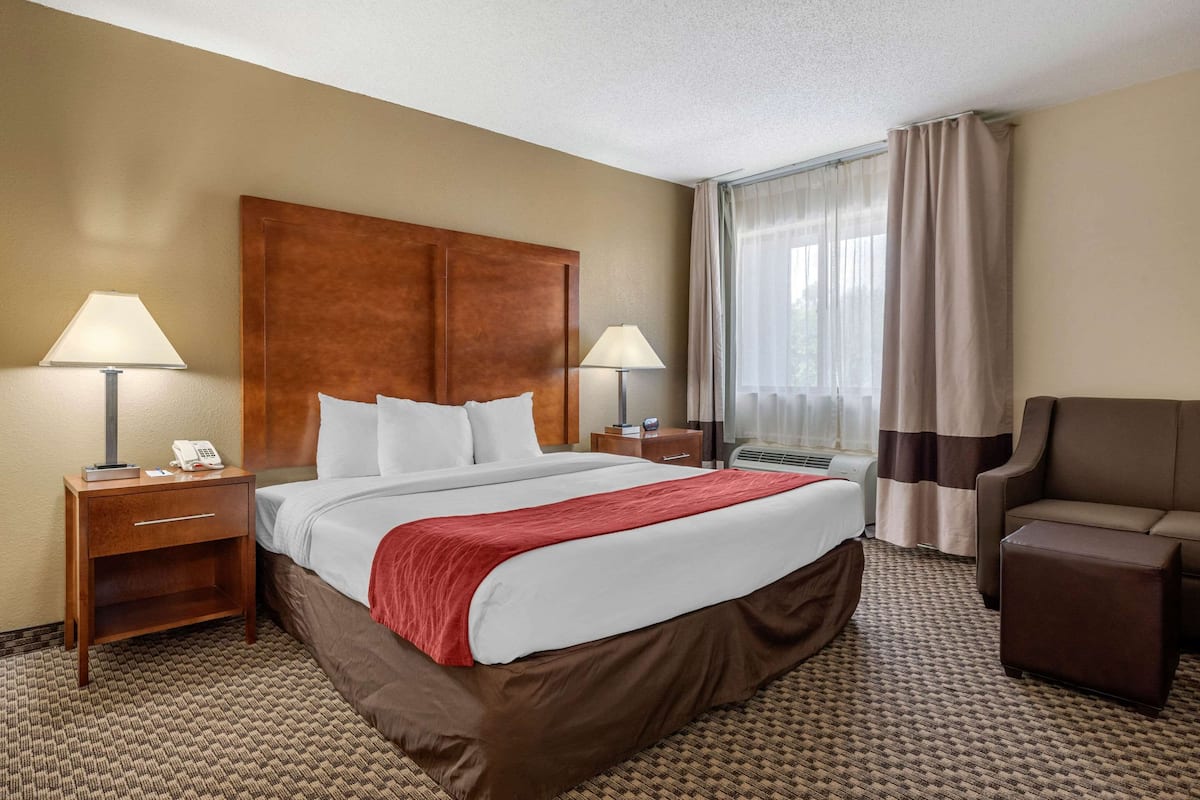 A hotel room with a large bed, two bedside tables with lamps, a brown armchair, and a window with curtains.