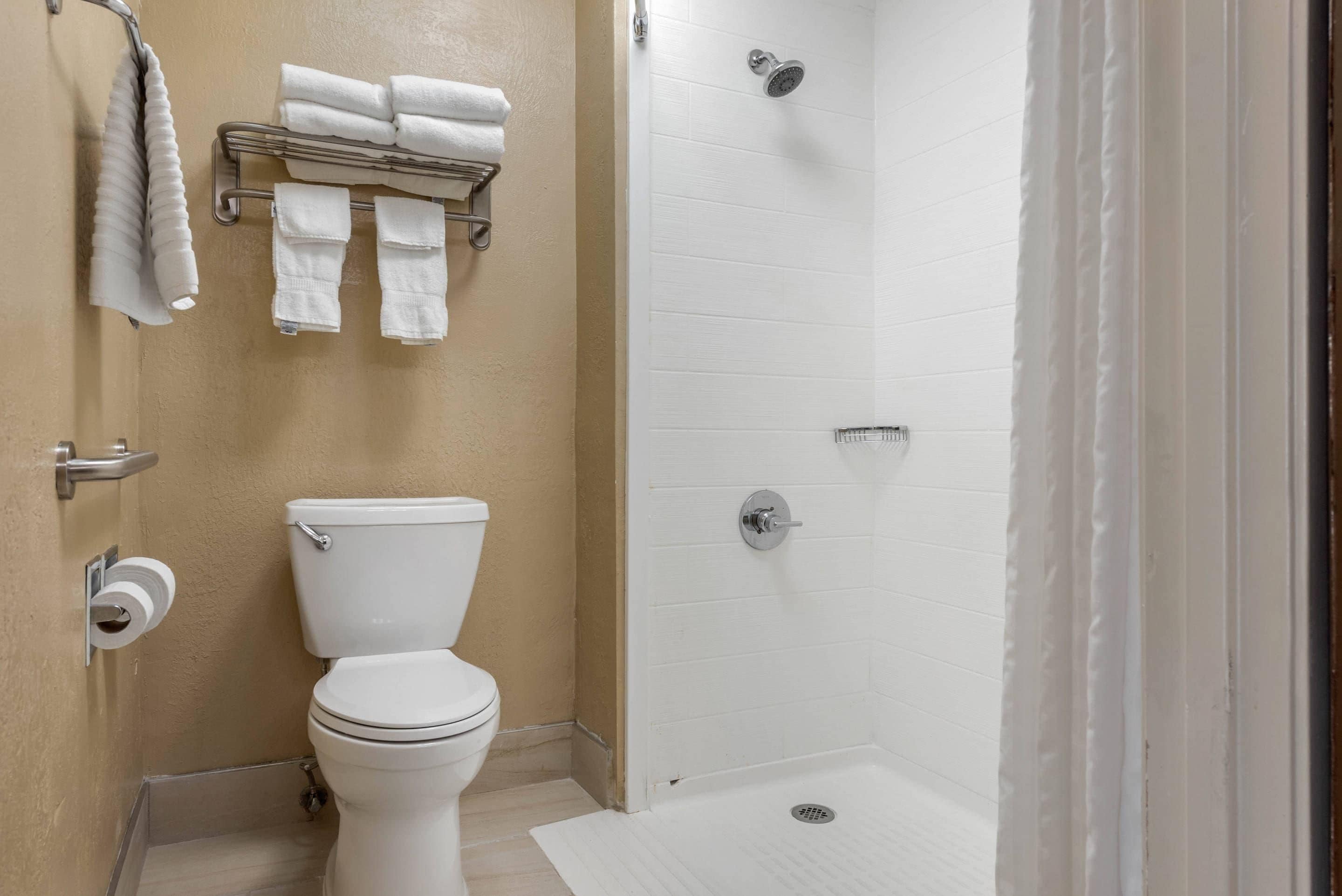 A bathroom with a toilet, a towel rack with folded towels, a showerhead, and a toilet paper holder.