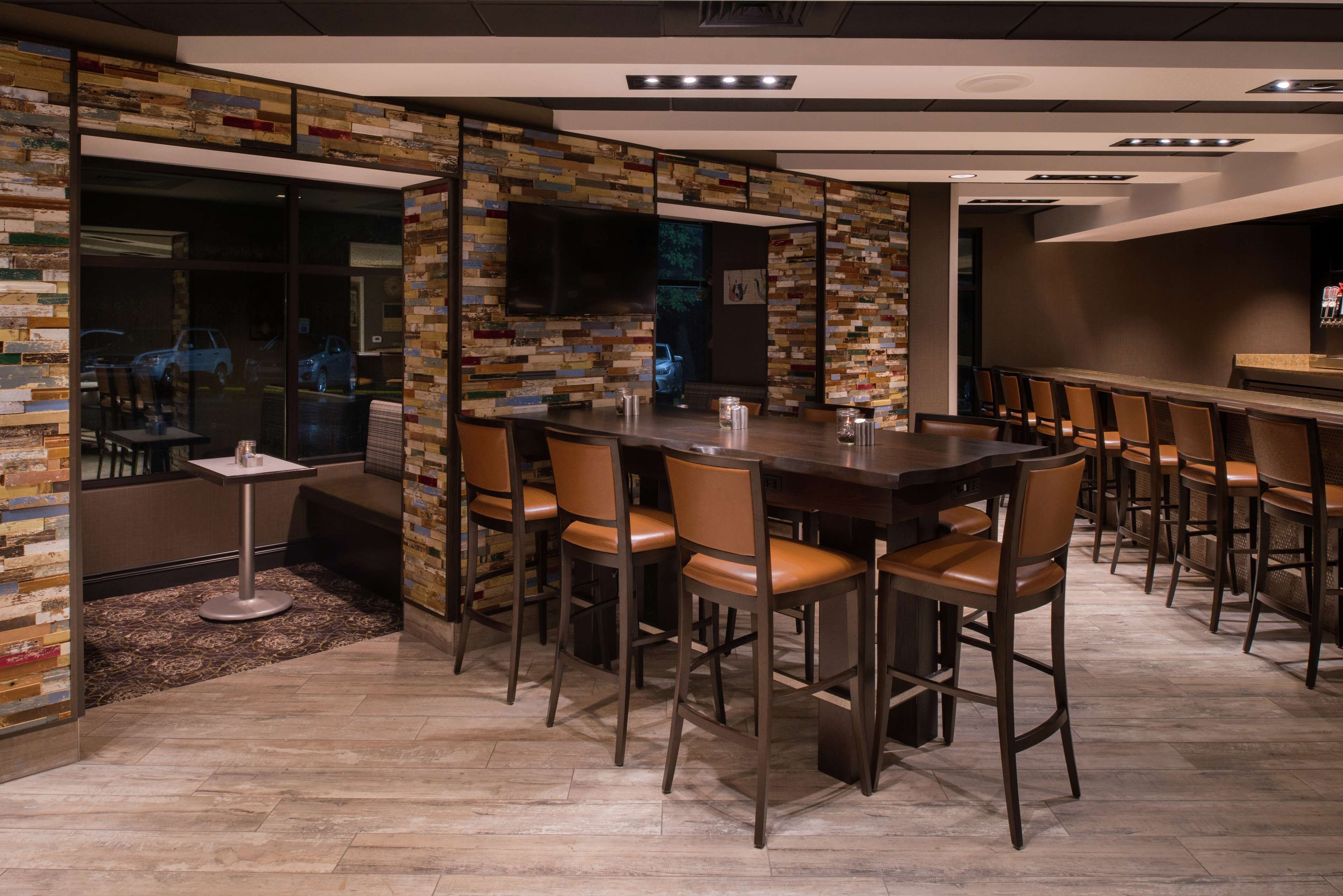 A modern bar area with a herringbone-patterned wall, a long wooden bar counter, and several leather chairs.