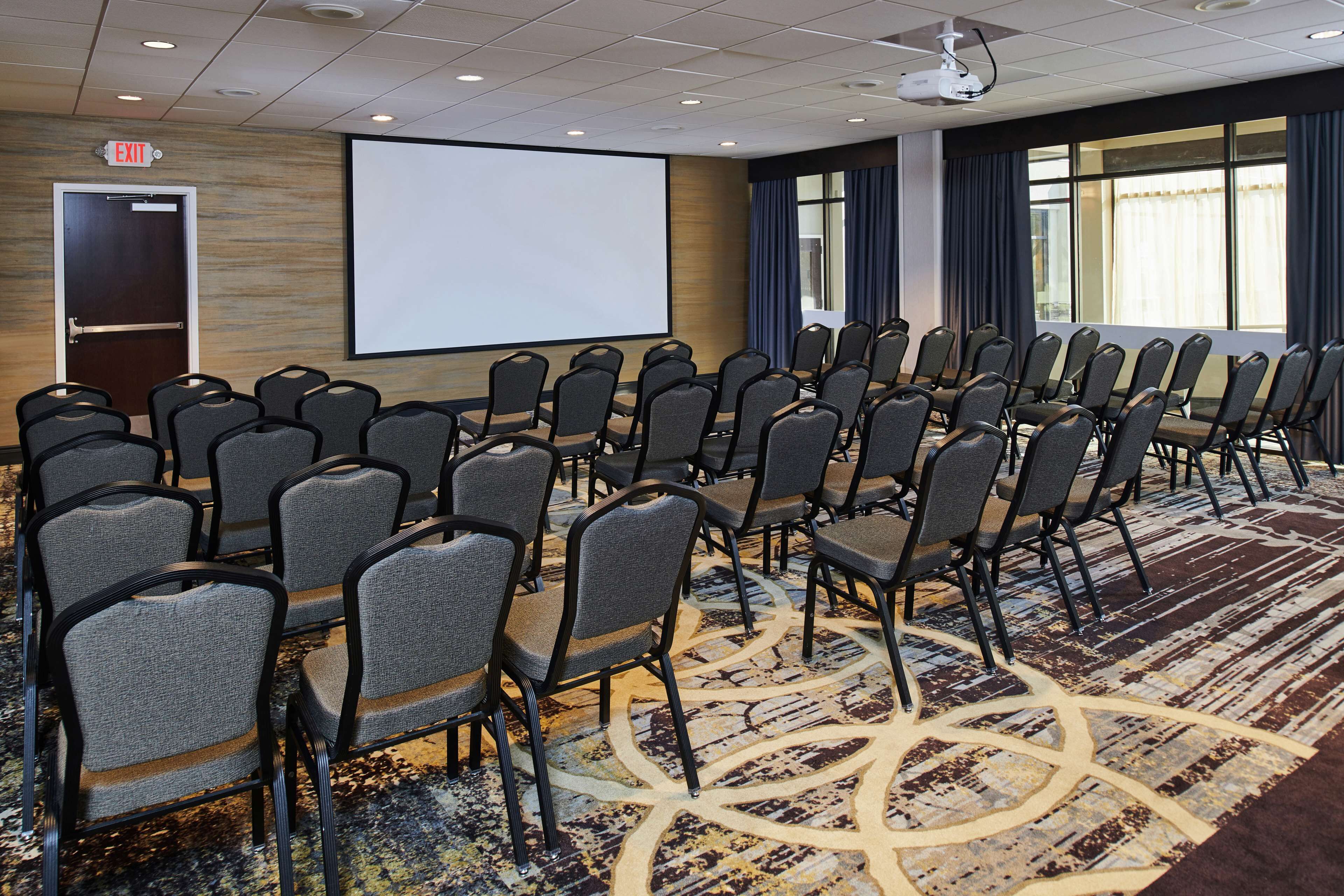 Une salle de conférence avec des rangées de chaises, un écran de projection et un panneau indiquant la sortie.
