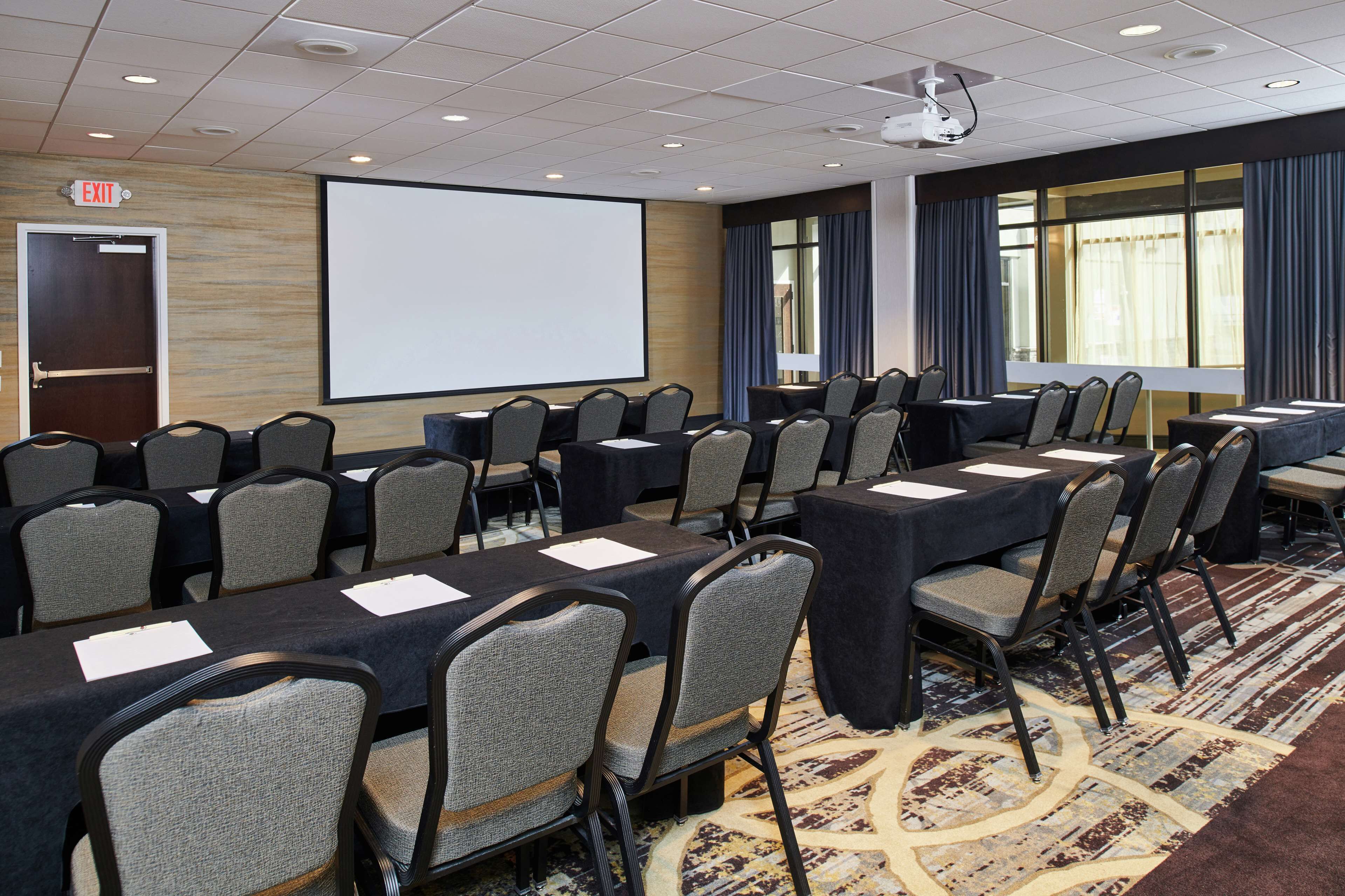 A conference room with multiple tables and chairs, a projector screen, and an exit door.