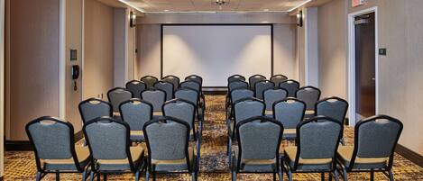 A conference room with rows of chairs facing a large screen, carpeted floor, and a ceiling with recessed lighting.