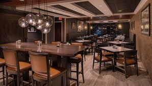 A restaurant with wooden tables and chairs, hanging pendant lights, and a brick wall. - DoubleTree by Hilton Raleigh Crabtree Valley (Raleigh)