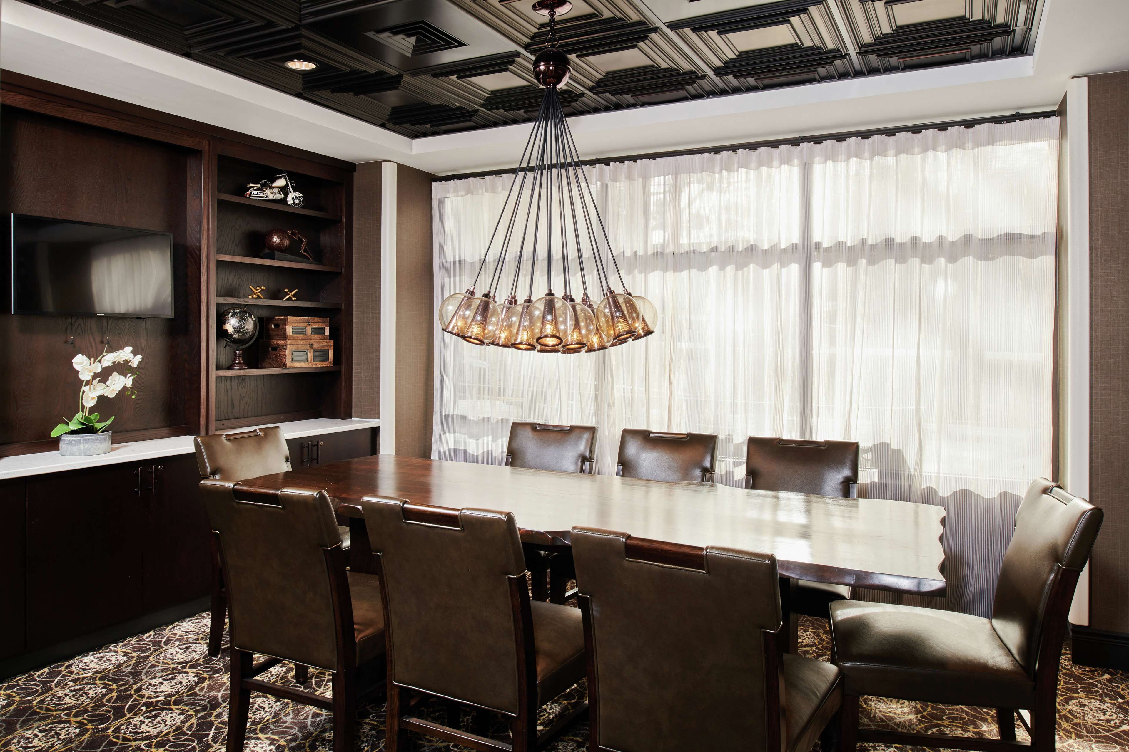 A dining area with a large wooden table, leather chairs, a chandelier, and a view through sheer curtains.