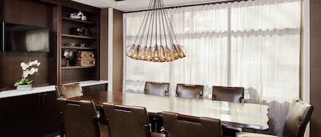 A dining area with a large wooden table, leather chairs, a chandelier, and a view through sheer curtains.