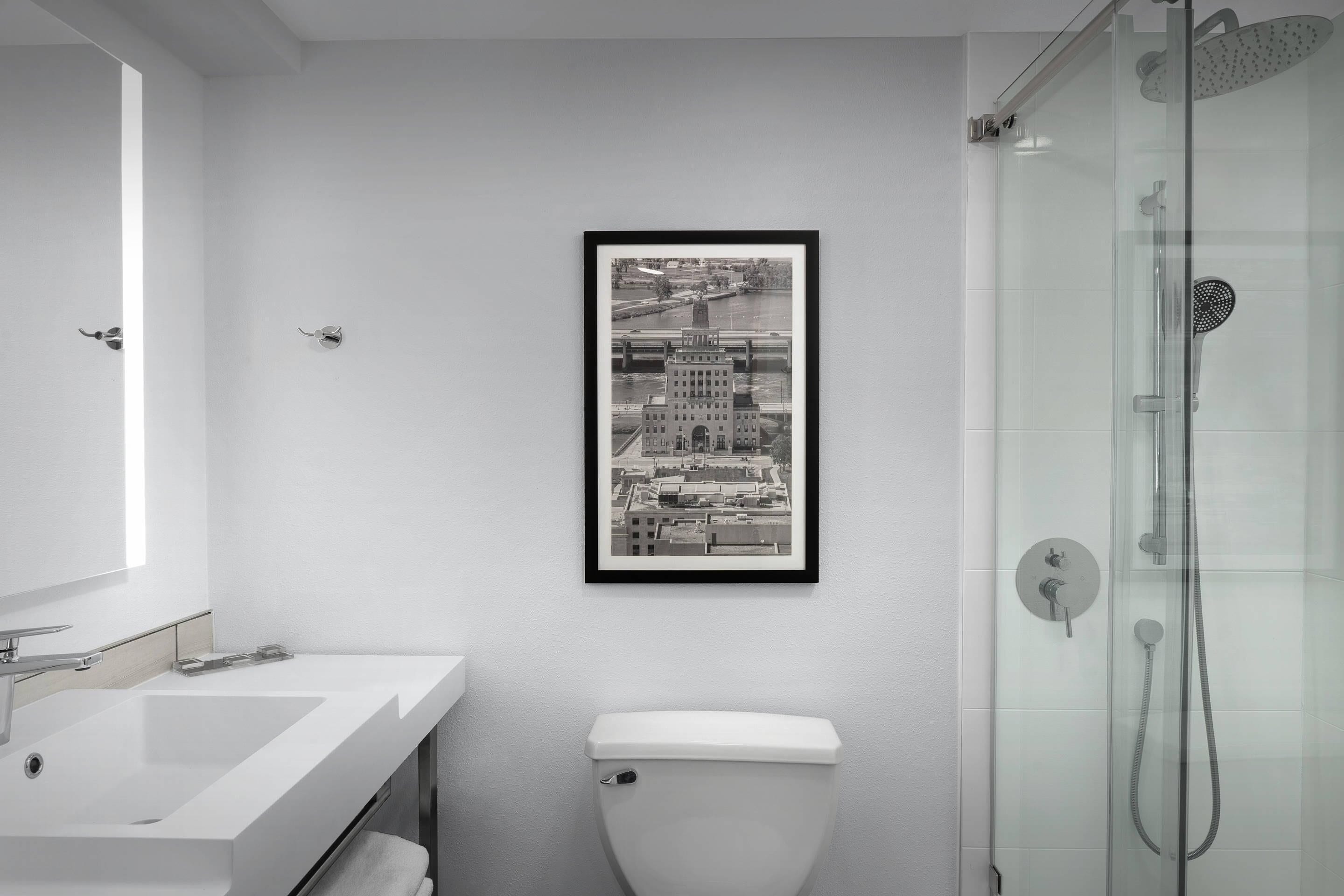 A modern bathroom with a white sink, toilet, and glass-enclosed shower. A framed cityscape photograph hangs on the wall.