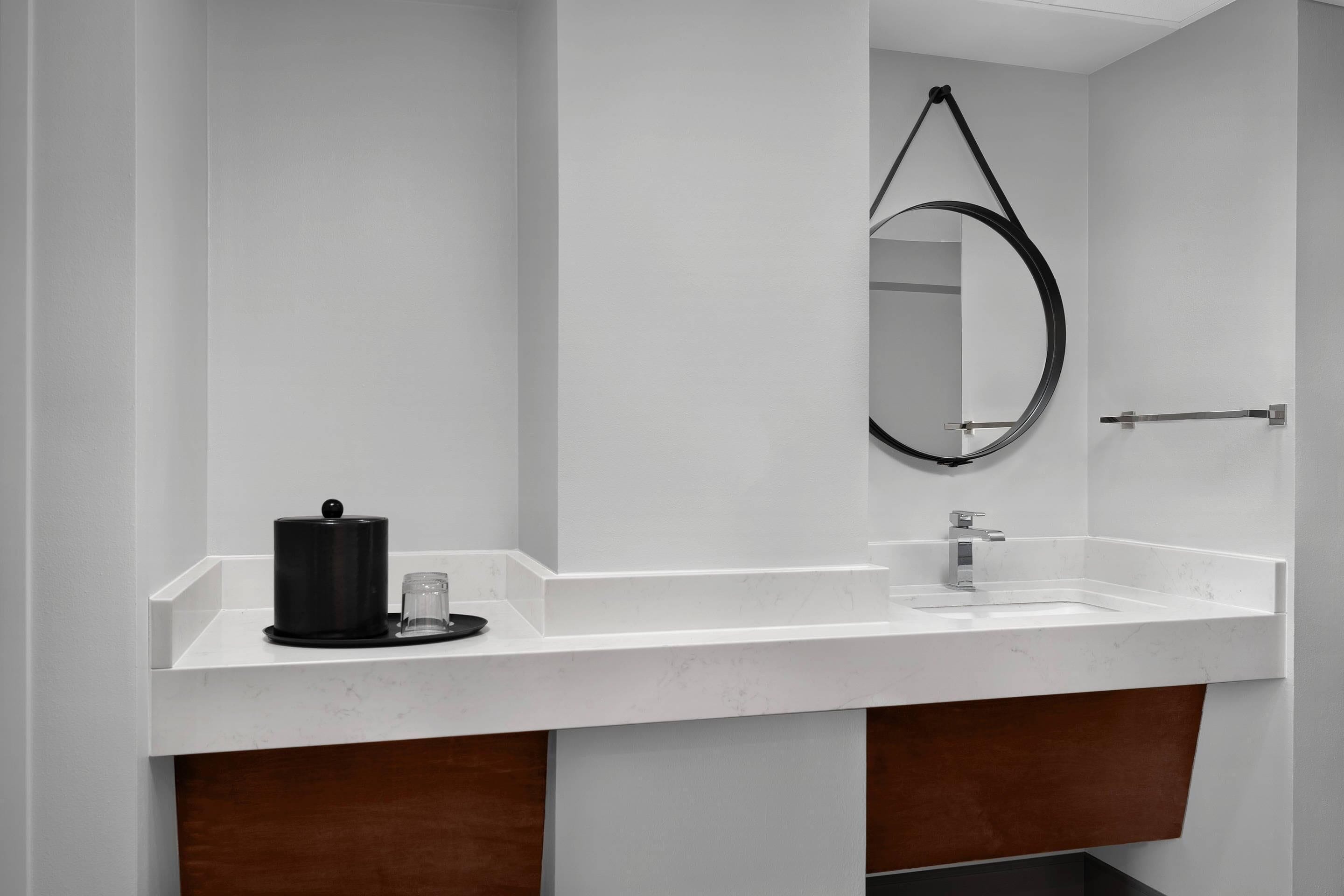 Een moderne badkamer met een witte wastafel, een zwarte koffiezetapparaat, een spiegel met zwarte lijst en een houten kastje onder de wastafel.