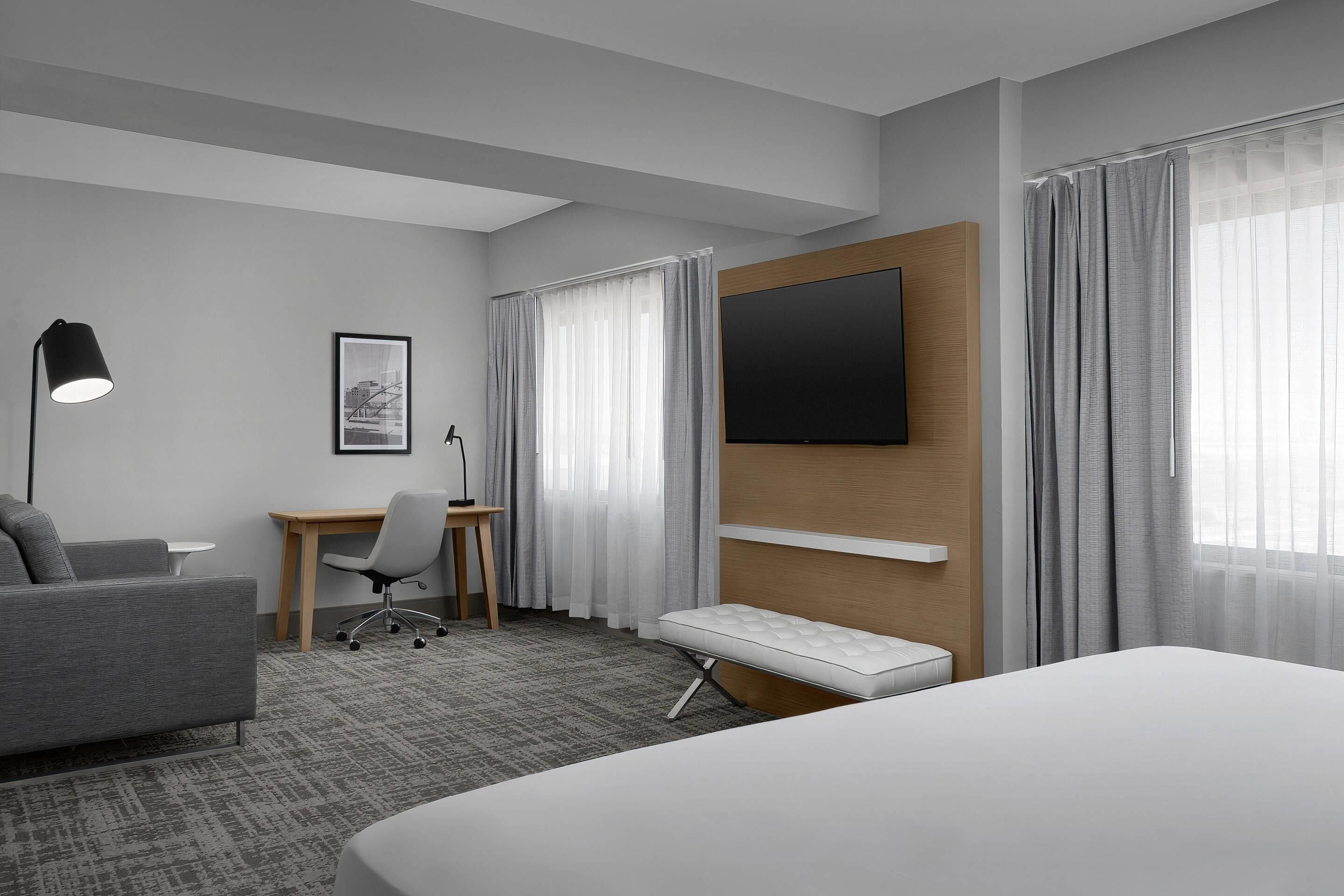 A hotel room with a bed, a desk, a chair, a lamp, a TV, and a window with curtains.